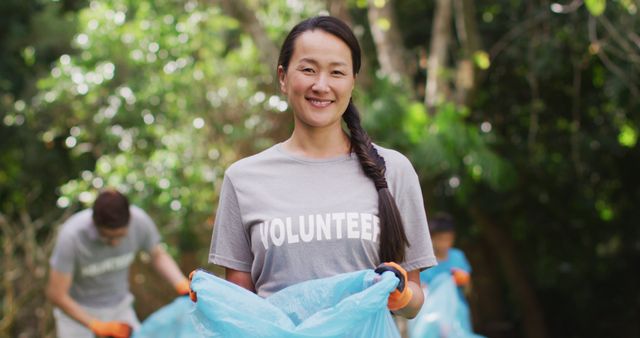 Smiling asian woman wearing volunteer t shirt holding refuse sack for collecting plastic waste. eco conservation volunteers doing countryside clean-up.