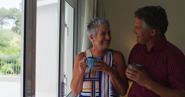 Middle-aged couple standing by a window at home, smiling and enjoying conversations while holding coffee cups. This can be used for themes around companionship, morning routines, relaxation, and lifestyle advertisements.
