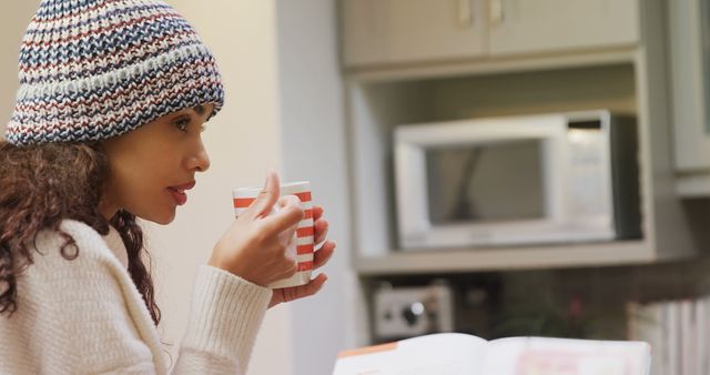 Woman wearing striped winter hat, enjoying hot beverage, likely tea or coffee, in kitchen. Book open on table. Ideal for articles, blogs, or advertisements about relaxation, winter comfort, or home lifestyle. Suitable for illustrating concepts of warmth, leisure, or casual indoor activities.