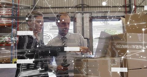 Warehouse managers collaborating with AR inventory analytics on laptop amid stacked boxes