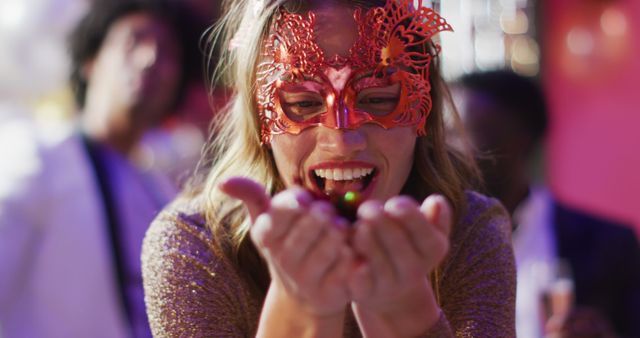 Image of happy caucasian woman in masquerade mask blowing glitter to camera at a nightclub. Fun, going out and party concept.