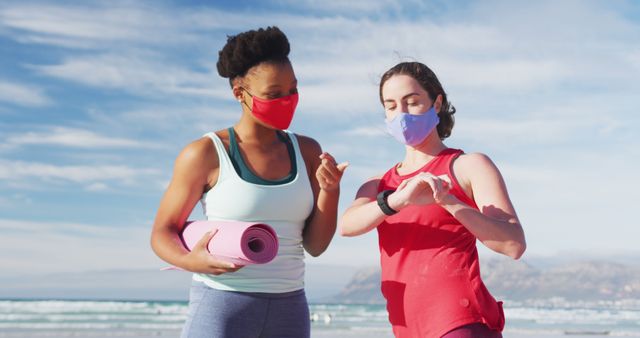 Two diverse female friends wearing face masks holding yoga mats at the beach. healthy active lifestyle, outdoor fitness and wellbeing.