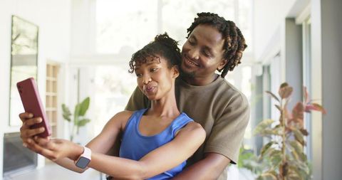 African american couple embracing while taking selfie, woman holding phone and wearing smartwatch