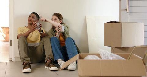 Young couple sitting on tiled floor with moving boxes holding mugs pointing toward new home