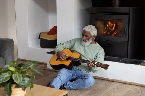 Senior african american man playing acoustic guitar by wood-burning stove in living room