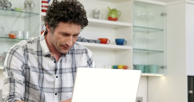 Middle-aged man using laptop in a bright, modern home kitchen. The kitchen features glass shelves displaying mugs and glassware. The man is focused and appears to be working or studying. This image is perfect for themes related to remote work, technology use at home, home offices, or general indoor activities.