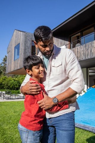 Father and young son sharing a heartwarming hug on grassy lawn in front of a contemporary wooden house with a balcony gives a sense of familial love and connection. The bright blue tent suggests an outdoor activity or adventure, symbolizing a relaxed environment. Ideal for themes related to family, togetherness, modern living, and outdoor experiences. Perfect for advertising family travel packages, outdoor gear, or home-related content.