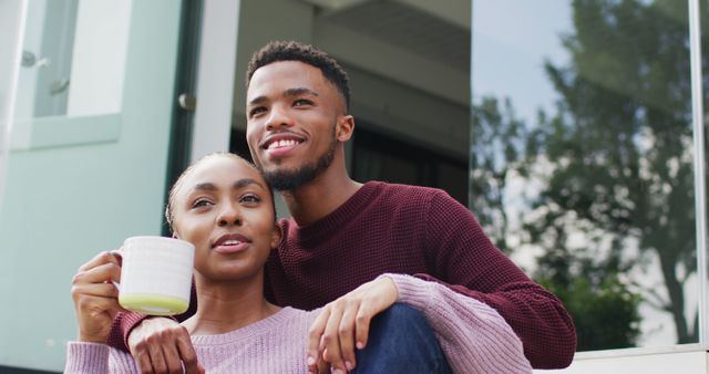 African American couple relaxing together outdoors with coffee, looking content and happy. Ideal for promotions on lifestyle, relationships, coffee brands, and outdoor leisure activities. Perfect for blogs or advertisements focusing on love, bonding, and morning routines.