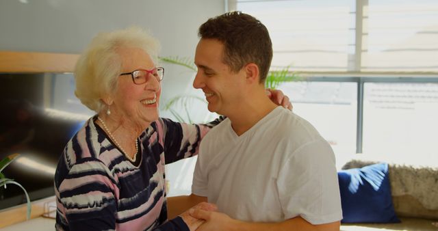 Elderly woman and her grandson sharing a joyful moment in a sunlit living room. Perfect for advertisements promoting family values, senior living, healthcare, or multigenerational households.