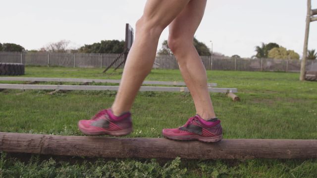 This depicts a young Caucasian woman skillfully balancing on a log as part of her workout in an outdoor gym setting, likely during a bootcamp session. With green grass in the background and her mid-stride motion captured perfectly, this can be used in fitness and exercise promotion for outdoor workout programs or agility and balance training tutorials.