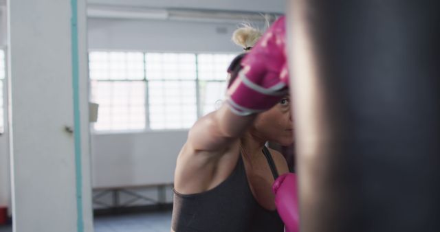 Woman practicing boxing by punching a heavy bag in a gym. She is wearing pink gloves, highlighting her focus and dedication to fitness and training. Ideal for illustrating themes of perseverance, female empowerment, and fitness routines.