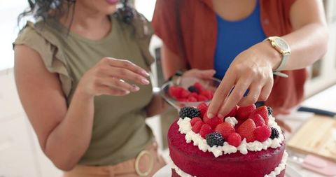 Diverse female friends decorating red velvet cake with raspberries and blackberries in sunlit kitche