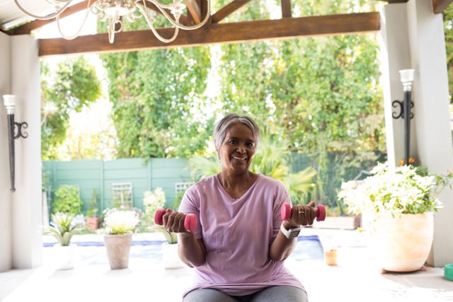 Senior woman sitting in yard, holding pink dumbbells, and smiling while exercising. Ideal for content related to fitness, healthy aging, home workouts, and promoting an active lifestyle among seniors. Can be used in articles, blogs, and advertisements focused on wellness and exercise for older adults.