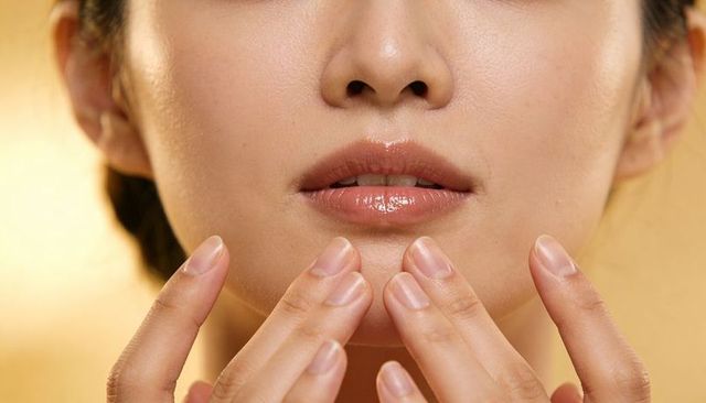 Asian woman touching chin with neat fingertips showcasing glossy lips and dewy skin