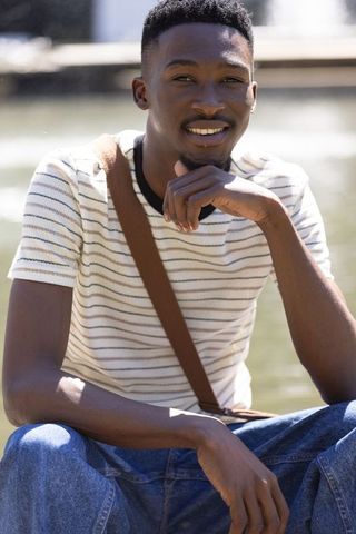 African american man sitting by pond smiling, wearing striped tee and brown crossbody bag