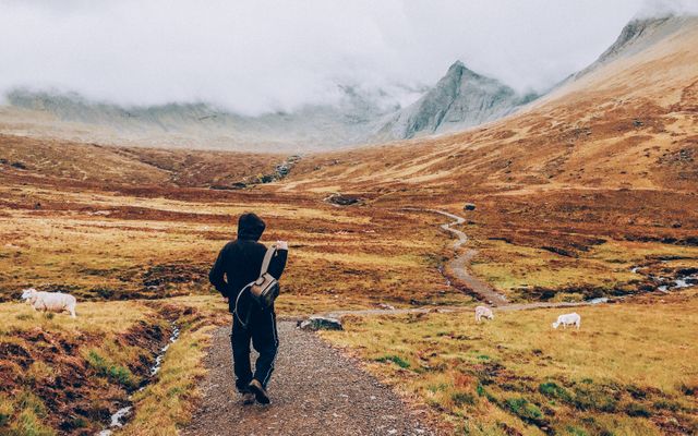 Man hiking a winding path through a mountainous rural landscape with cloudy skies. This image captures the essence of adventure, solitude, and exploring nature's beauty. Ideal for use in travel brochures, adventure blogs, or outdoor lifestyle promotions.