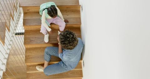 Mother helping daughter with sneaker on wooden stairs indoor