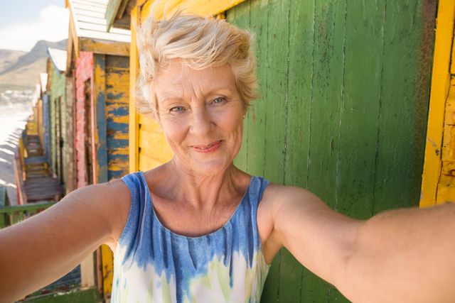 Senior woman smiling and taking a selfie in front of colorful beach huts on a sunny day. Ideal for use in travel brochures, lifestyle blogs, retirement community advertisements, and summer vacation promotions.