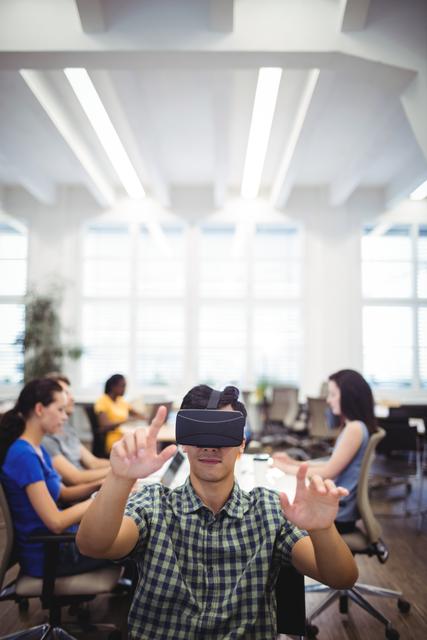 Man using virtual reality headset in a contemporary open office layout. His coworkers are seen working in the background. This image can be used for depicting themes of technological advancement, innovation in workplaces, VR applications in business, and modern office environments.