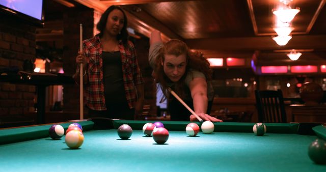 Two women are engaged in a game of pool in a dimly lit billiards room, with copy space. One woman, focused and leaning over the table, takes her shot while the other observes patiently.
