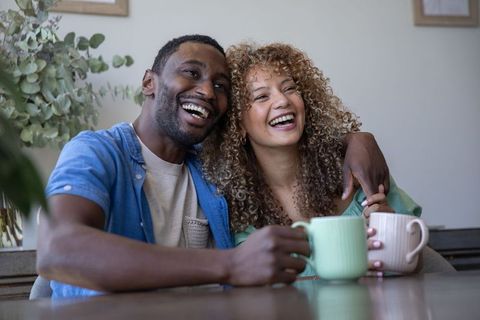 Interracial couple laughing and holding mugs while sitting at cozy dining table