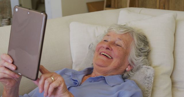 Elderly woman is lying on sofa, engaging with her tablet, and enjoying herself. The woman seems happy and relaxed, using modern technology to stay connected or entertained. Suitable for illustrating tech-savvy seniors, promoting digital literacy among older individuals, and depicting joyful moments in everyday life.