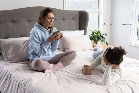 Cozy morning chat between diverse female friends on bed holding coffee mugs
