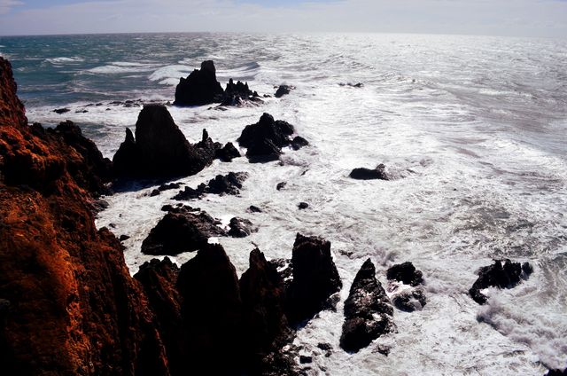 Dramatic seascape capturing waves crashing against jagged black rocks, emphasizing the power and roughness of the ocean. Rust-colored cliffs contrast sharply with the turbulent white waters. Ideal for creating atmospheric scenes, emphasizing nature's power, or for backgrounds needing rugged coastal scenery.