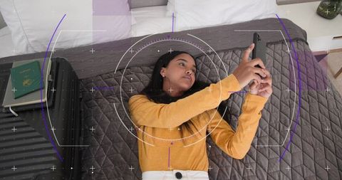 Woman lying on bed scrolling smartphone with suitcase and passport, travel hud overlay
