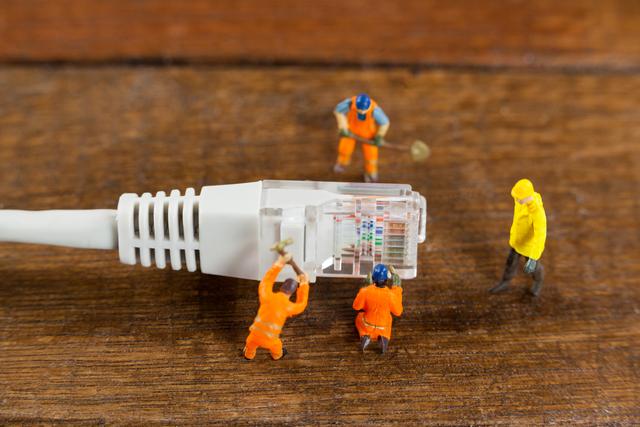 Image depicts small figures of workers fixing a LAN cable on a wooden surface, symbolizing technology and teamwork. Ideal for technology-related content, network maintenance illustrations, or articles on internet connectivity and repair. Perfect for blogs, advertisements, and educational materials.