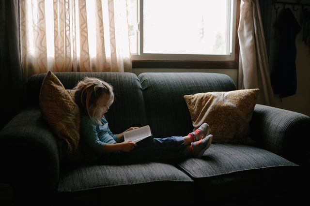 Little girl is lying on a couch reading a book in the soft glow of natural window light. The setting exudes a peaceful and cozy atmosphere with two decorative pillows. Can be used for educational content, children's ebook covers, or articles on childhood relaxation and indoor activities.