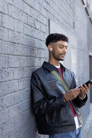 Asian young man listening to earbuds while checking smartphone against urban brick wall