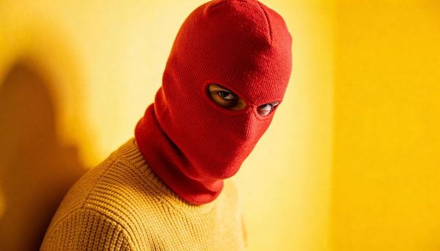Male-presenting portrait wearing red balaclava and mustard sweater on yellow backdrop