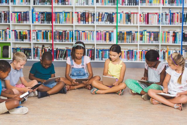 This image shows a diverse group of elementary school children sitting on the floor of a library, using digital tablets. The background features bookshelves filled with books, indicating a learning environment. This image can be used for educational websites, school brochures, technology in education articles, and promotional materials for libraries or educational programs.