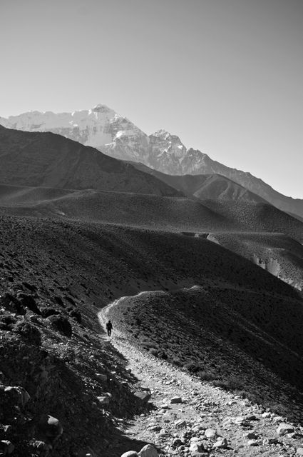 This photograph captures a lone hiker walking on a rugged mountain trail in a vast, remote landscape. The monochromatic treatment emphasizes the stark beauty of the rugged terrain and towering peaks. Ideal for use in travel blogs, adventure magazines, outdoor gear advertisements, and motivational posters.