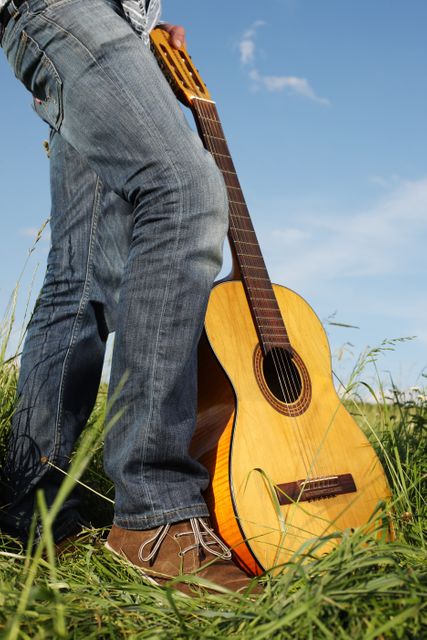 Man holding a guitar while standing in a grassy field, wearing jeans and boots. Suitable for themes of music, relaxation, nature, country lifestyle, creativity, or personal leisure. Great for use in advertisements, social media posts, blog articles, or website banners associated with outdoor activities or music-related content.