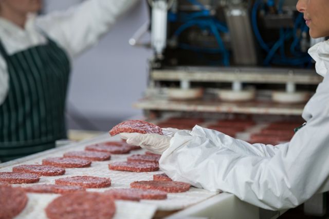 Female butcher wearing gloves inspecting hamburger patties on conveyor belt at meat processing plant. Image useful for themes related to food manufacturing, quality control, industrial work, and food hygiene. Suitable for articles and materials discussing meat production, factory operations, and workplace safety.