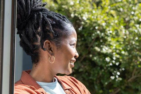 African woman smiling looking toward sunlit garden in rust jacket with gold hoop earrings
