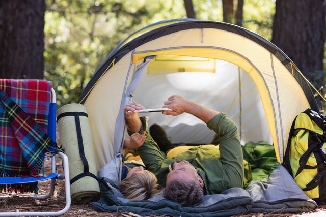 Father and son lying inside a tent at a campsite, using a digital tablet. Surrounded by nature, they are enjoying a bonding moment in the forest. Ideal for use in articles or advertisements about family vacations, outdoor activities, camping gear, or technology in nature.