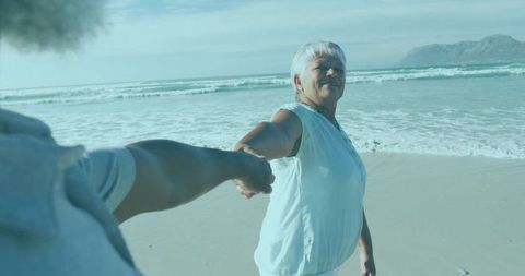 Senior woman holding hand on sunlit beach, smiling and enjoying seaside stroll