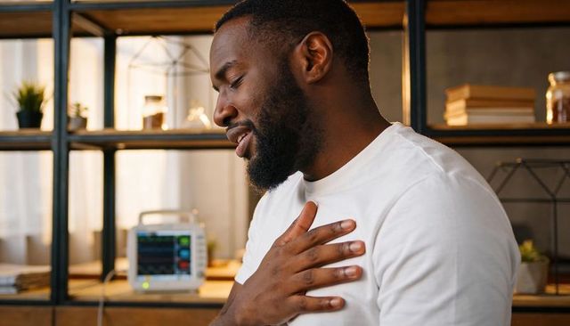 Black man holding hand to chest in home office near medical monitor showing heartbeat