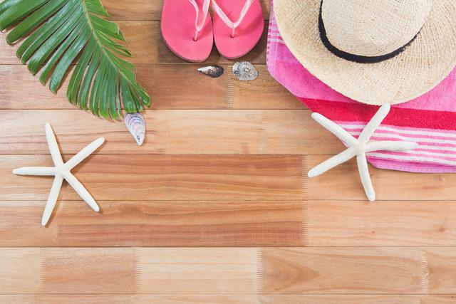 Beach accessories including a straw hat, pink flip flops, starfish, seashells, a pink towel, and a green leaf on a wooden board. Perfect for use in travel blogs, vacation promotions, summer event advertisements, and beach resort commercials.