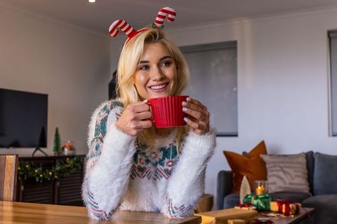 Smiling woman holding red mug wearing candy cane headband enjoying cozy holiday morning
