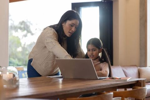 Mother teaching daughter on laptop at dining table for remote learning and homework help