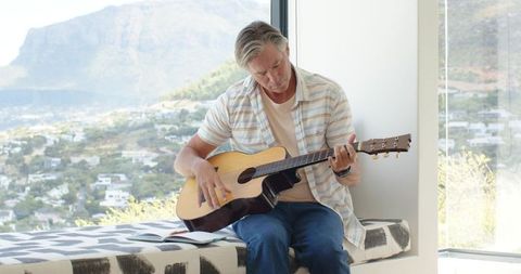 Mature man playing acoustic guitar by window with hillside view, sunlit minimal interior