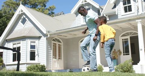 African american father and son playing soccer on sunny front lawn of white suburban home