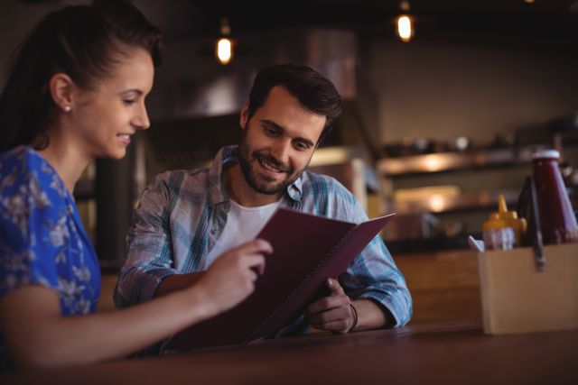 Couple sitting at a table in a cozy bar, reviewing the menu together. They are smiling and appear to be enjoying their time. This image can be used for promoting restaurants, bars, dining experiences, date night ideas, and lifestyle blogs.