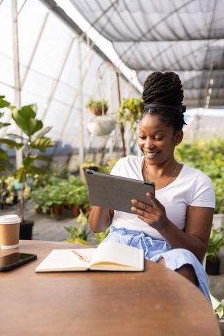 African american woman smiling using tablet in greenhouse among plants with notebook