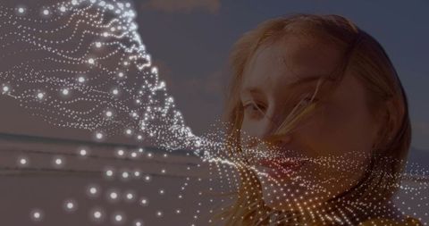 Adult woman letting wind move hair with glowing dotted particle mesh over sunset beach