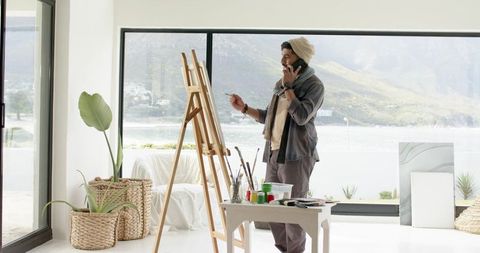 Indian artist painting in sunlit lakeside studio, holding phone, minimalist atelier